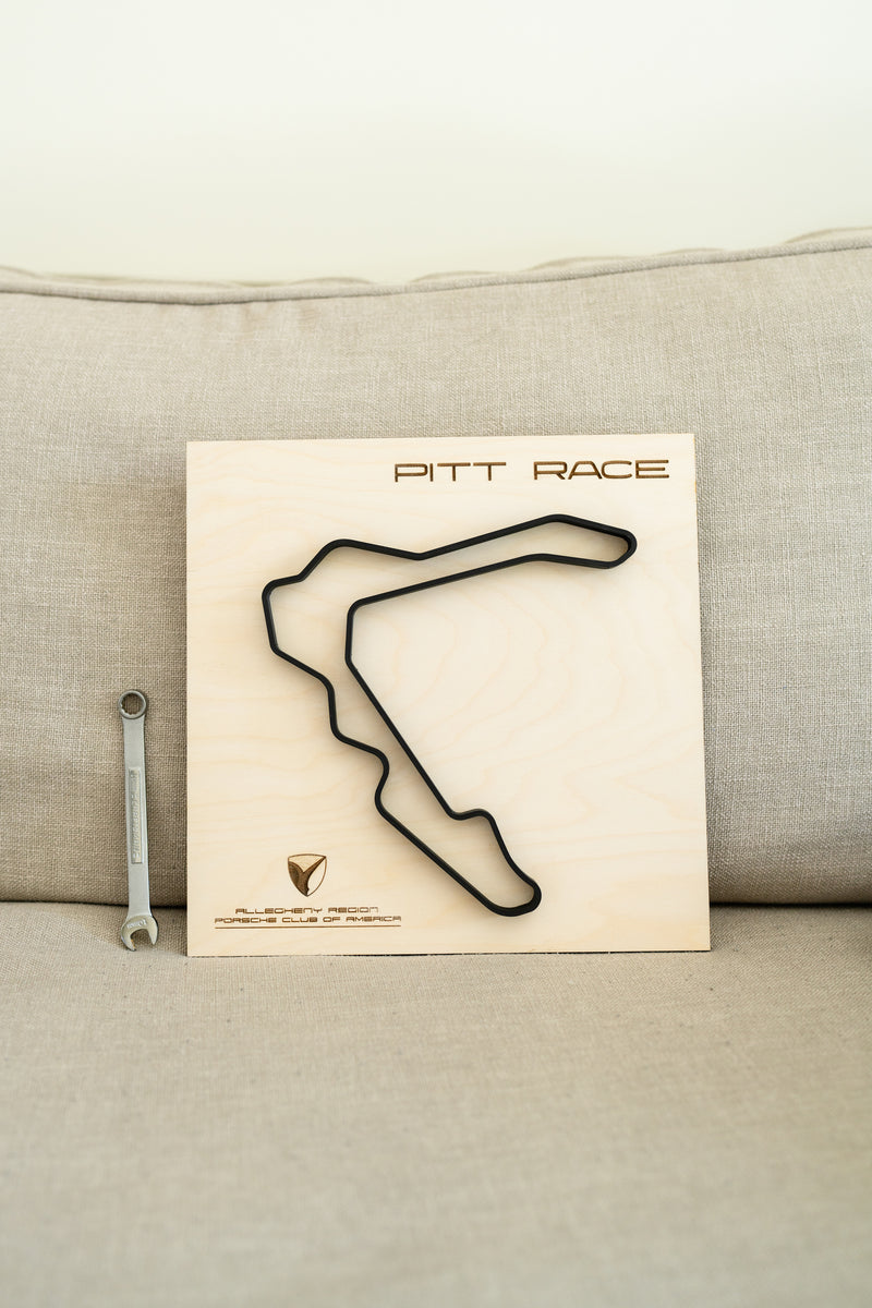 The product is resting on a sofa next to a wrench for scale. The product is a wood board with Pitt Race laser etched on the top. The Allegheny Region Porsche Club of America logo is laser etched on the bottom right. The Pitt Race track is centered on the wood board.